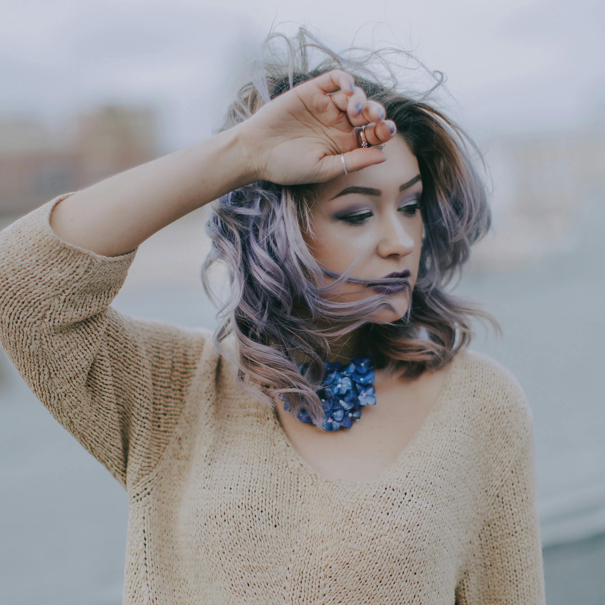 Fashionable woman with lilac hair and blue necklace posing outdoors on a windy day.